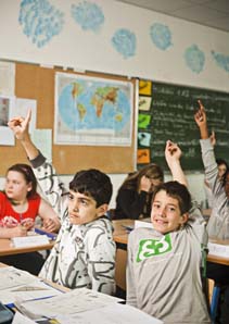Drei Kinder im Klassenzimmer zeigen mit den Fingern auf, andere  schauen nach vorne oder beugen sich über ihre Hefte