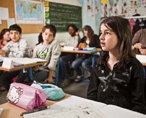 Ein Mädchen im Klassenzimmer schaut nach vorne sagt etwas, die  anderen Kinder blicken sie an