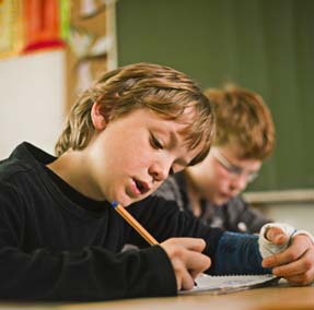 Ein Junge mit Gipsarm sitz an einem Pult im Klassenzimmer und  schreibt etwas
